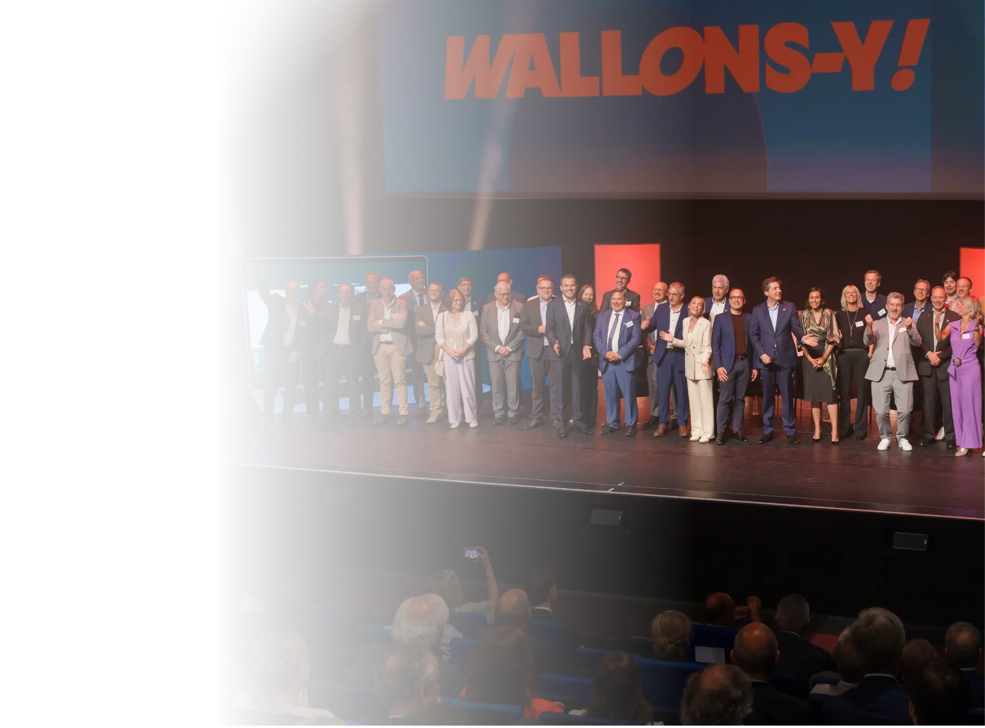 Un groupe de personnes habillées en tenue formelle se tient sur une scène devant un public avec un grand écran affichant le texte "WALLONS-Y !" en arrière-plan.

Description générée par IA