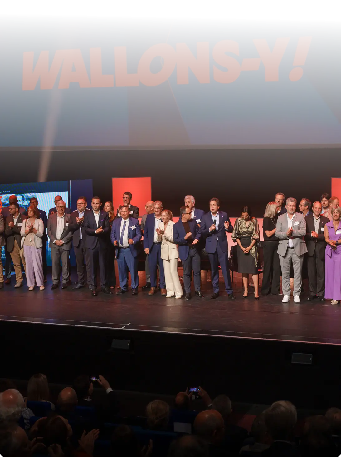 Un groupe de personnes habillées de manière formelle se tient sur une scène devant un écran affichant le texte "WALLONS-Y !" dans une salle de spectacle ou de conférence.

Description générée par IA