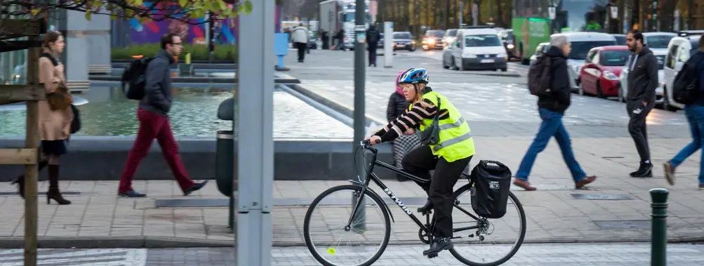 Une personne portant un gilet jaune réfléchissant et un casque fait du vélo sur une route urbaine tandis que plusieurs personnes marchent sur le trottoir à proximité.

Description générée par IA