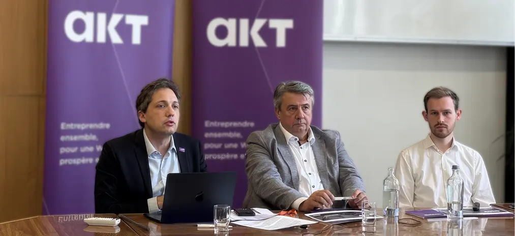 Trois adultes assis à une table en bois lors d'une réunion professionnelle avec des documents, un ordinateur portable et des bouteilles d'eau devant eux, avec deux panneaux violets portant le logo "akt" en arrière-plan.

Description générée par IA