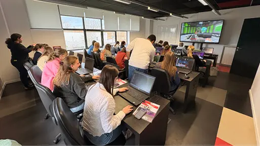 Des personnes assises dans une salle de classe moderne utilisent des ordinateurs portables tandis qu'une personne debout les aide, avec un grand écran affichant un jeu vidéo à l'avant de la pièce.

Description générée par IA