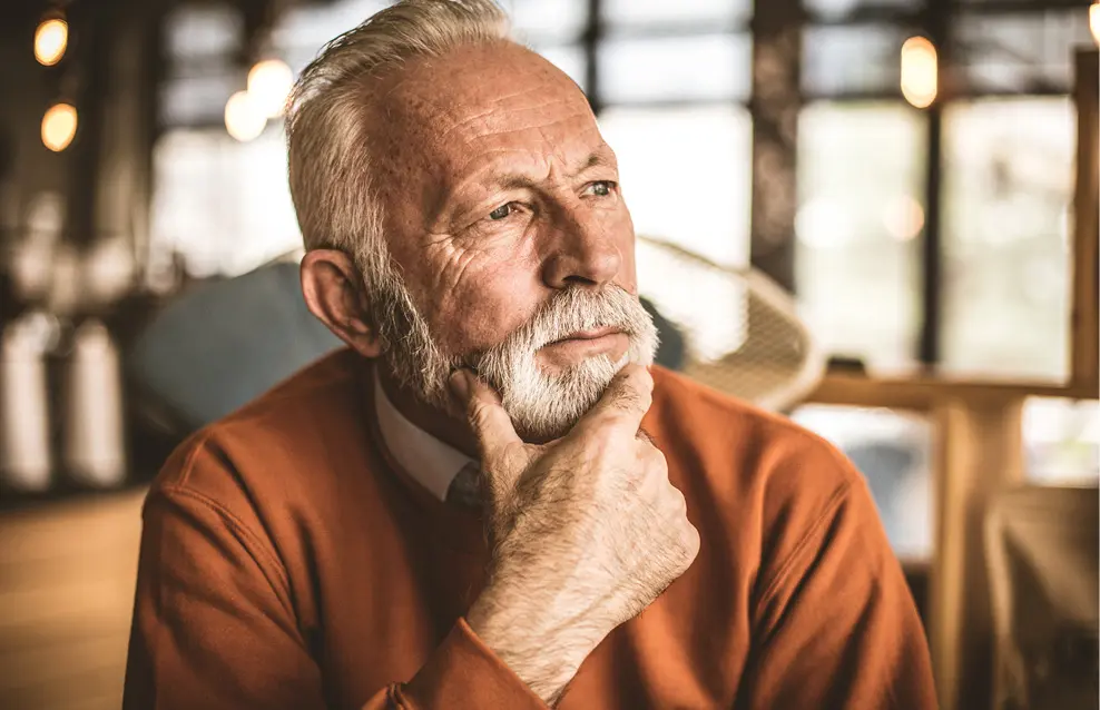 Une personne âgée portant un pull orange est assise dans un café lumineux en posant la main sur le menton dans une posture pensive.

Description générée par IA