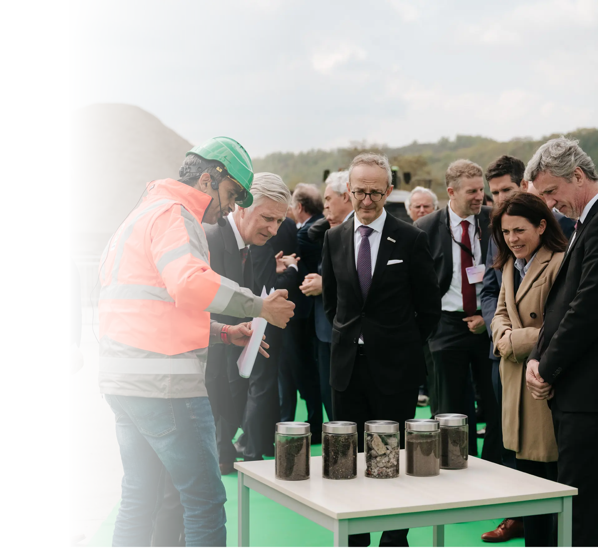 Un groupe de personnes en tenue professionnelle écoute un adulte en veste de sécurité et casque vert qui présente plusieurs bocaux contenant différents matériaux sur une table dans un environnement extérieur.

Description générée par IA