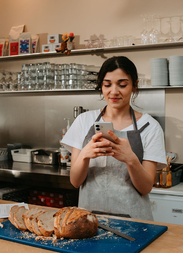 Une personne portant un tablier gris et un t-shirt blanc tient un téléphone portable dans une cuisine avec du pain tranché sur une planche bleue devant elle.

Description générée par IA