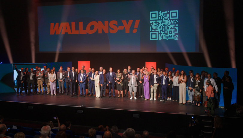 Un grand groupe de personnes habillées de manière formelle se tient sur une scène devant un écran affichant le texte "WALLONS-Y !" et un code QR dans une salle de spectacle avec un public assis.

Description générée par IA