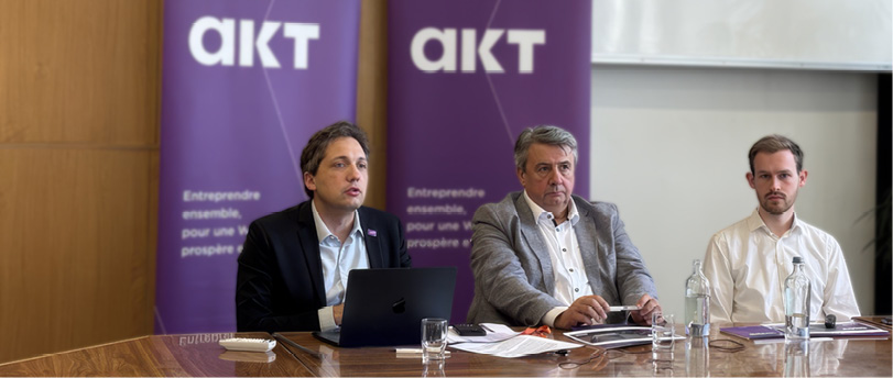 Trois adultes assis à une table en bois avec des documents, un ordinateur portable et des verres d'eau devant deux panneaux violets portant le logo "aikt".

Description générée par IA