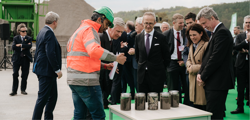 Un groupe de personnes en tenue professionnelle discute avec une personne en combinaison de travail orange et casque vert sur un chantier extérieur.

Description générée par IA