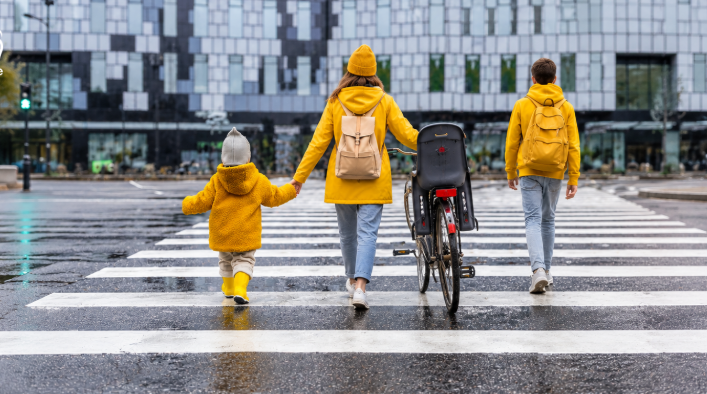 Trois personnes portant des vêtements jaunes traversent un passage piéton sous la pluie, dont une pousse un vélo.

Description générée par IA