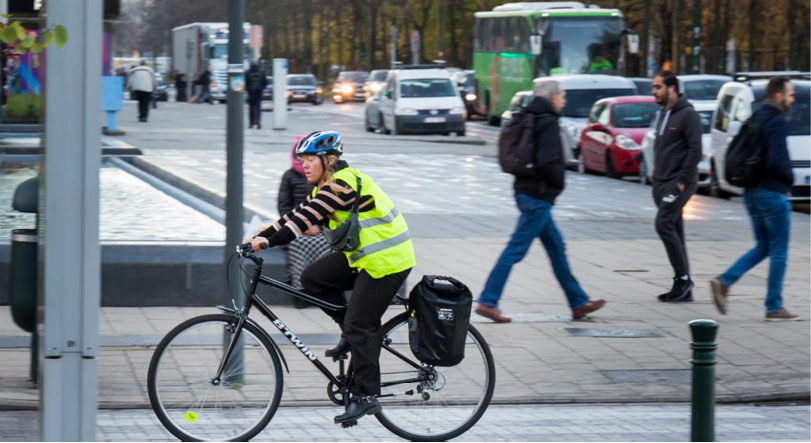 Un adulte portant un gilet jaune réfléchissant et un casque fait du vélo sur une piste cyclable en ville tandis que plusieurs personnes marchent sur le trottoir à proximité.

Description générée par IA