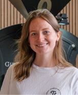 Une personne aux cheveux longs porte un t-shirt blanc avec un logo circulaire et se tient devant une machine industrielle noire avec un fond en bois vertical.

Description générée par IA