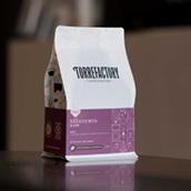 Un paquet de café blanc avec le logo "TORREFACTORY Coffee Roasters" et une étiquette violette indiquant "Milamores" posé sur une surface floue en arrière-plan.

Description générée par IA
