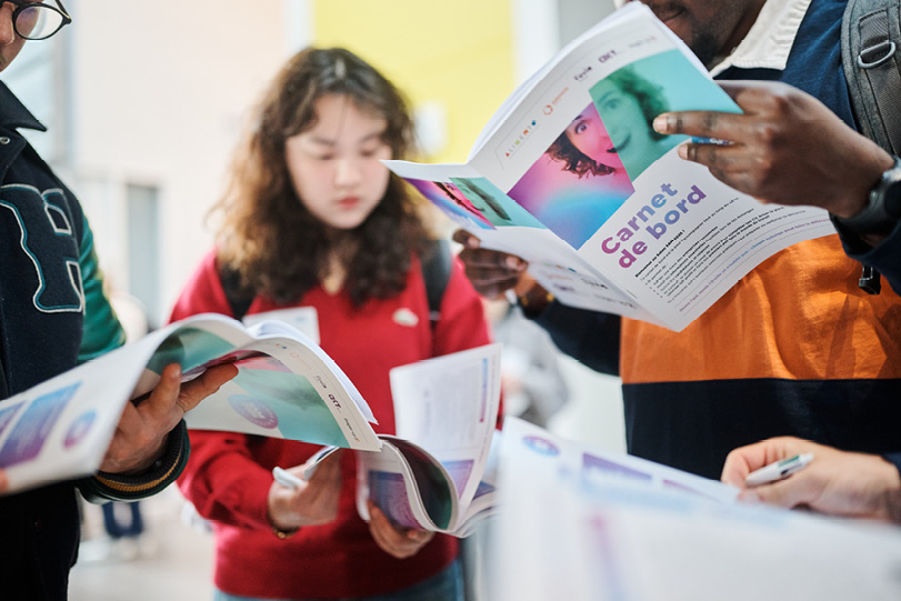 Des personnes debout lisant des brochures colorées dans un environnement intérieur lumineux.

Description générée par IA