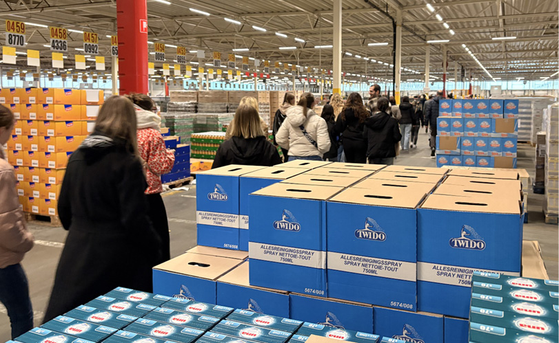 Des personnes vêtues de manteaux marchent dans un grand entrepôt rempli de palettes de cartons empilés sous un plafond métallique éclairé.

Description générée par IA