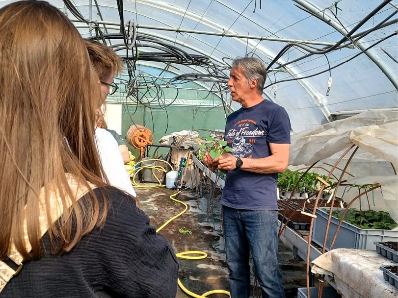 Des personnes discutent dans une serre entourées de plantes et de matériel de jardinage sous une structure en plastique transparent.

Description générée par IA